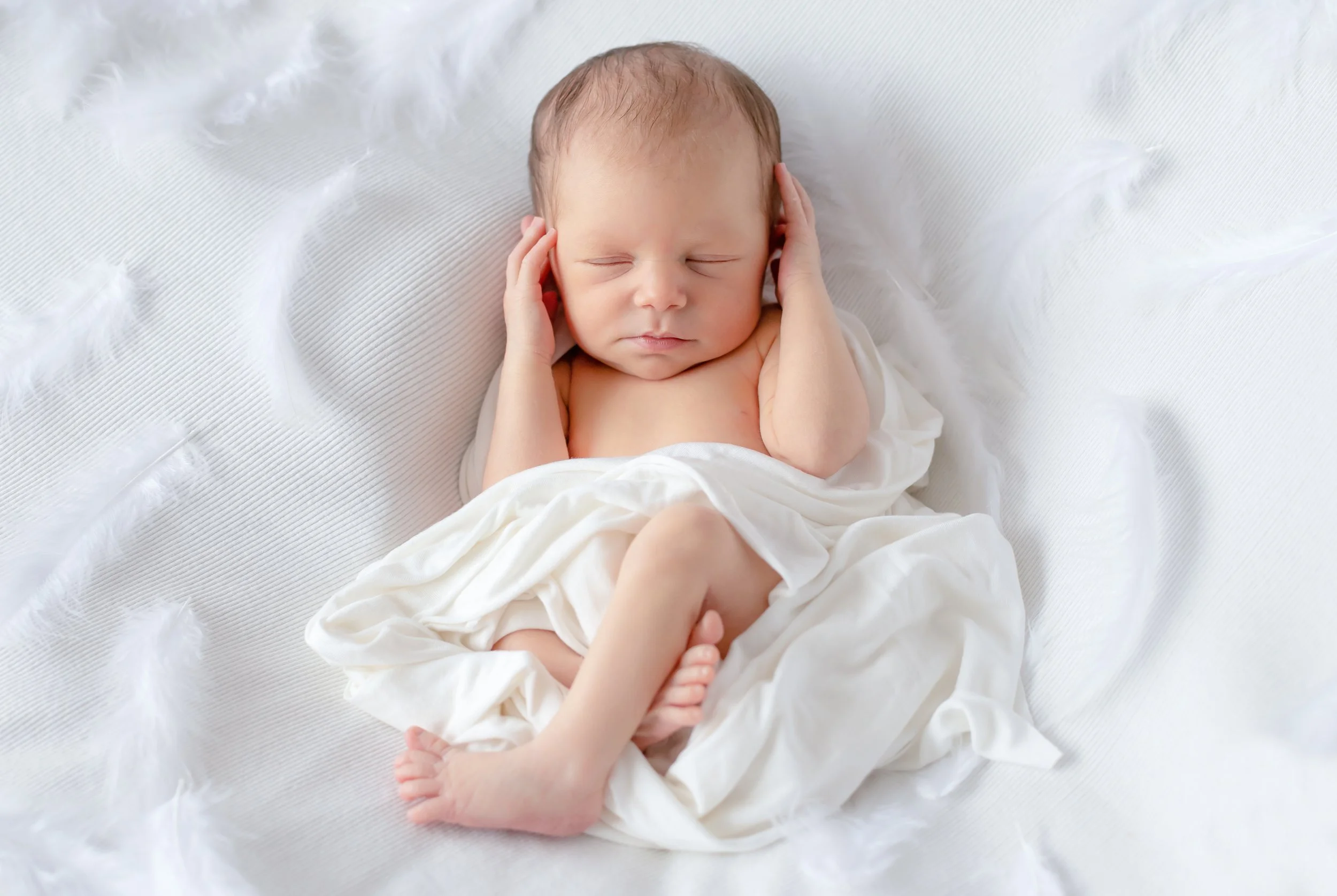 A newborn baby with closed eyes lying on a white textured surface, wrapped in white cloth with fluffy feather details nearby.