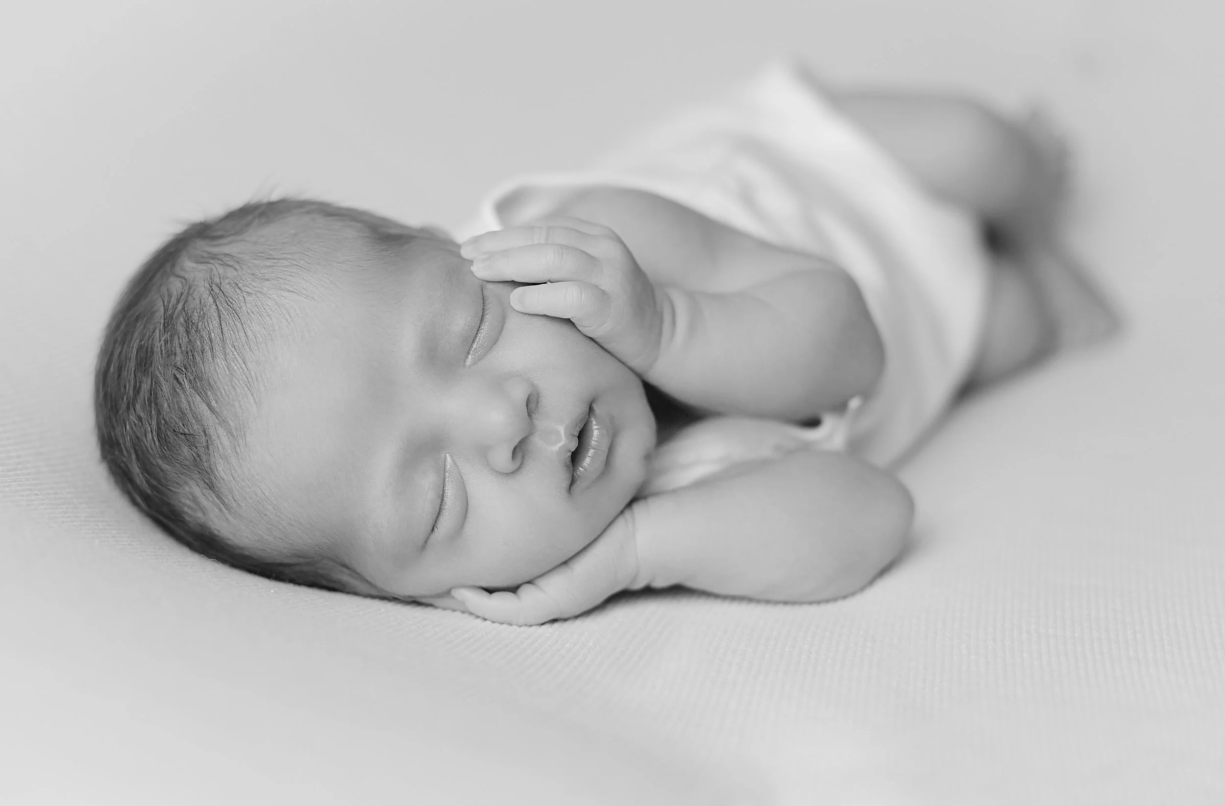 Black and white photo of a sleeping newborn baby with hand on forehead, lying on a soft surface.