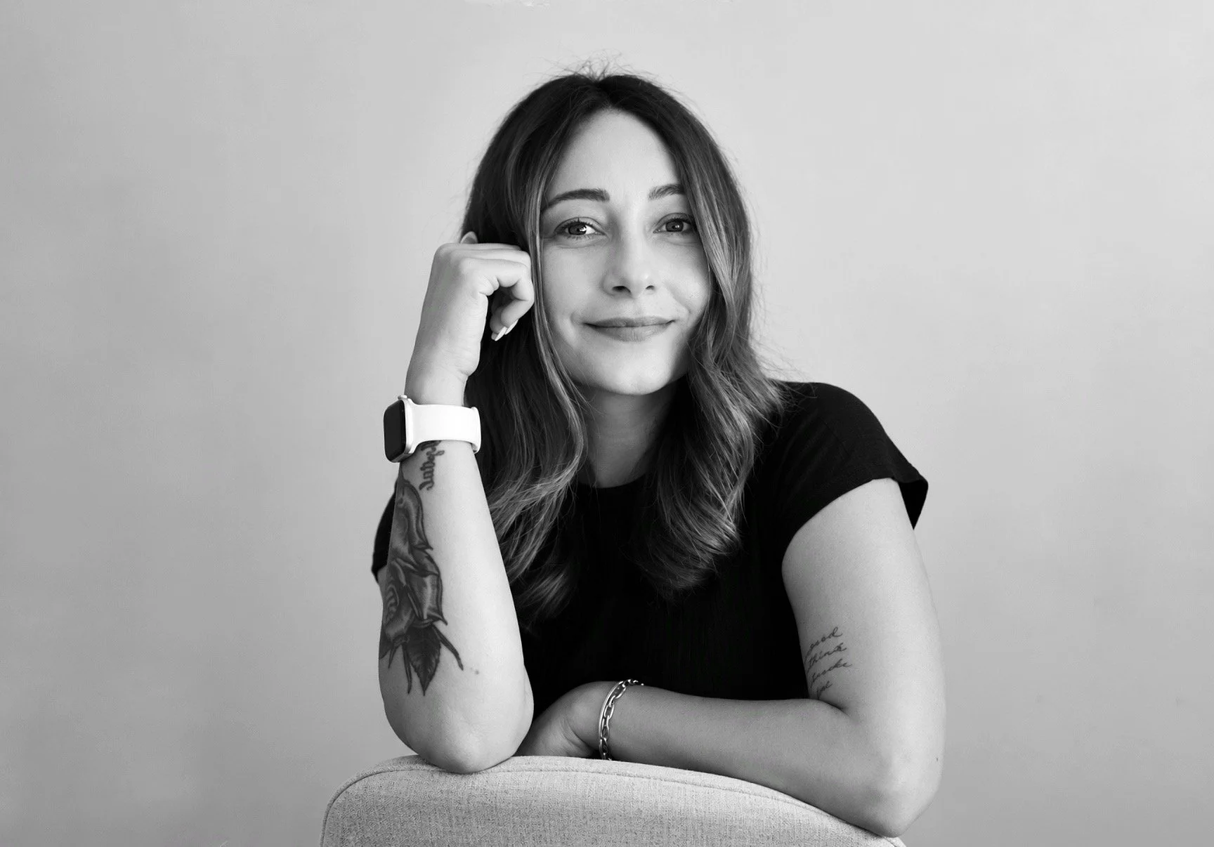 Black and white portrait of a smiling woman with long wavy hair, resting her head on her hand, wearing a black shirt, a wristwatch, and a bracelet, sitting in front of a plain light background.