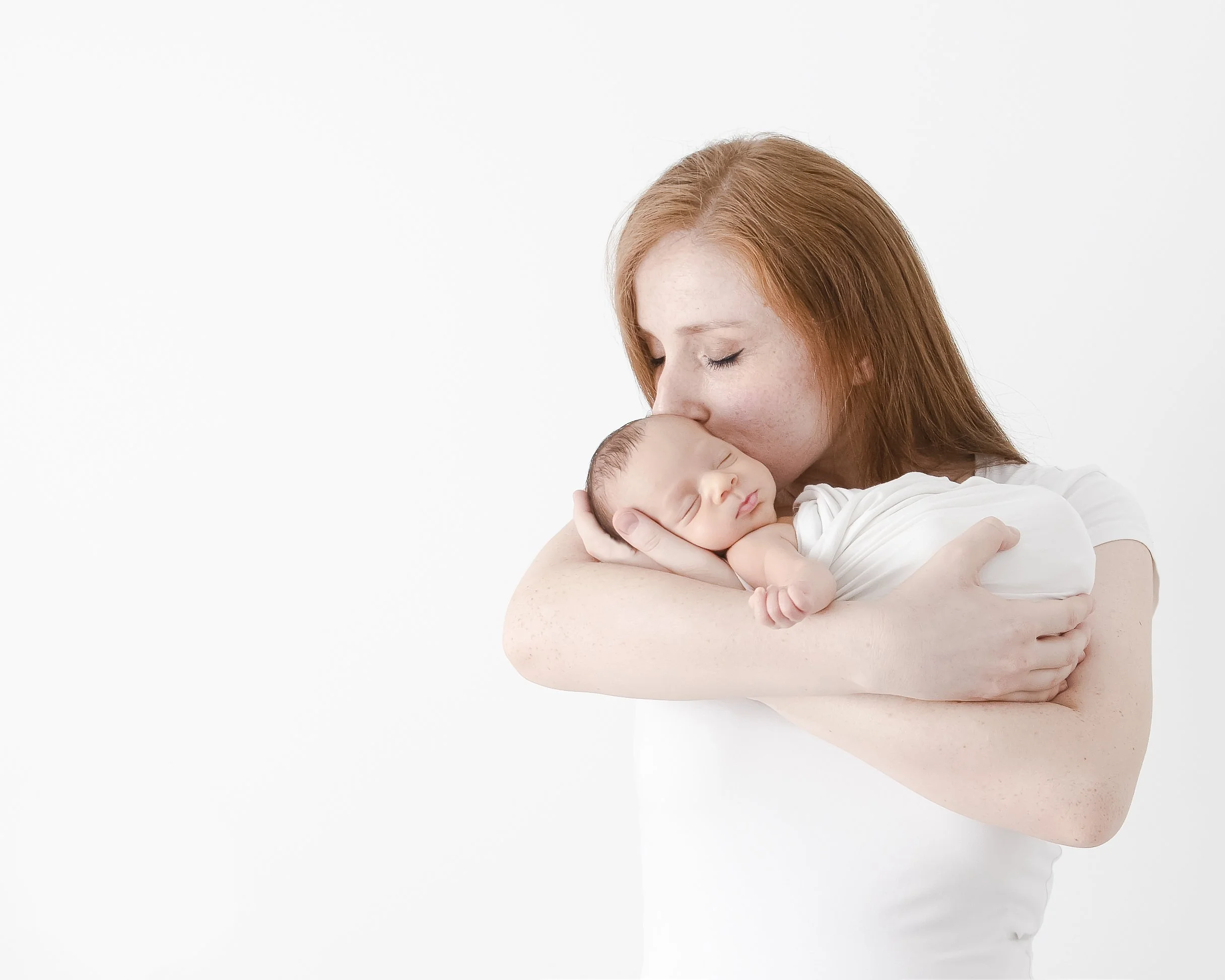 A woman with red hair tenderly holds a sleeping newborn baby close to her face, both against a plain white background.
