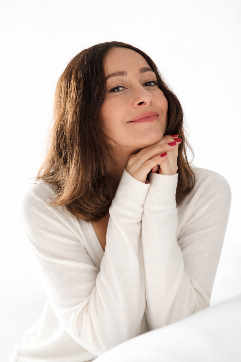 A woman with shoulder-length brown hair, smiling softly with her hands under her chin, wearing a white top, against a white background.
