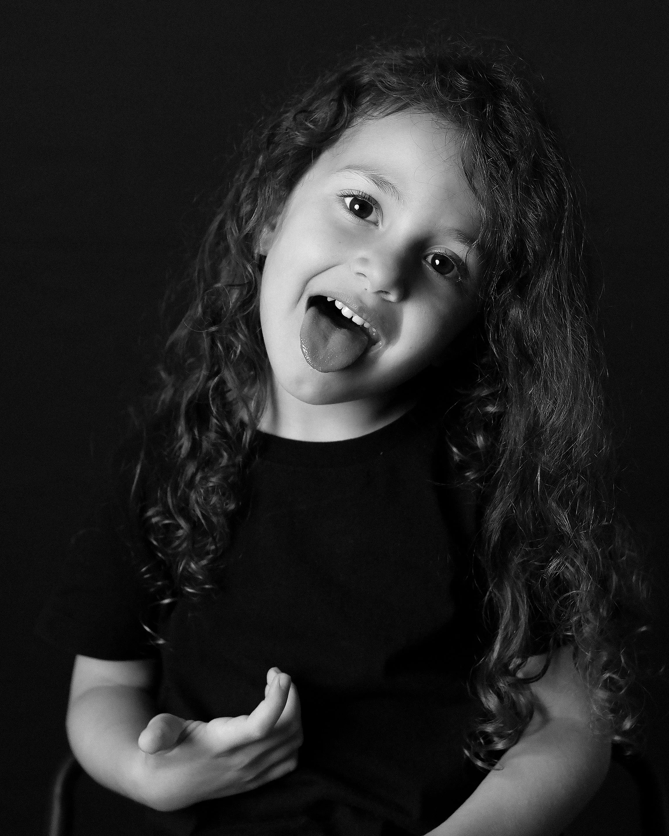 A young girl with curly hair making a funny face with her tongue sticking out and her eye wide open, wearing a black shirt.