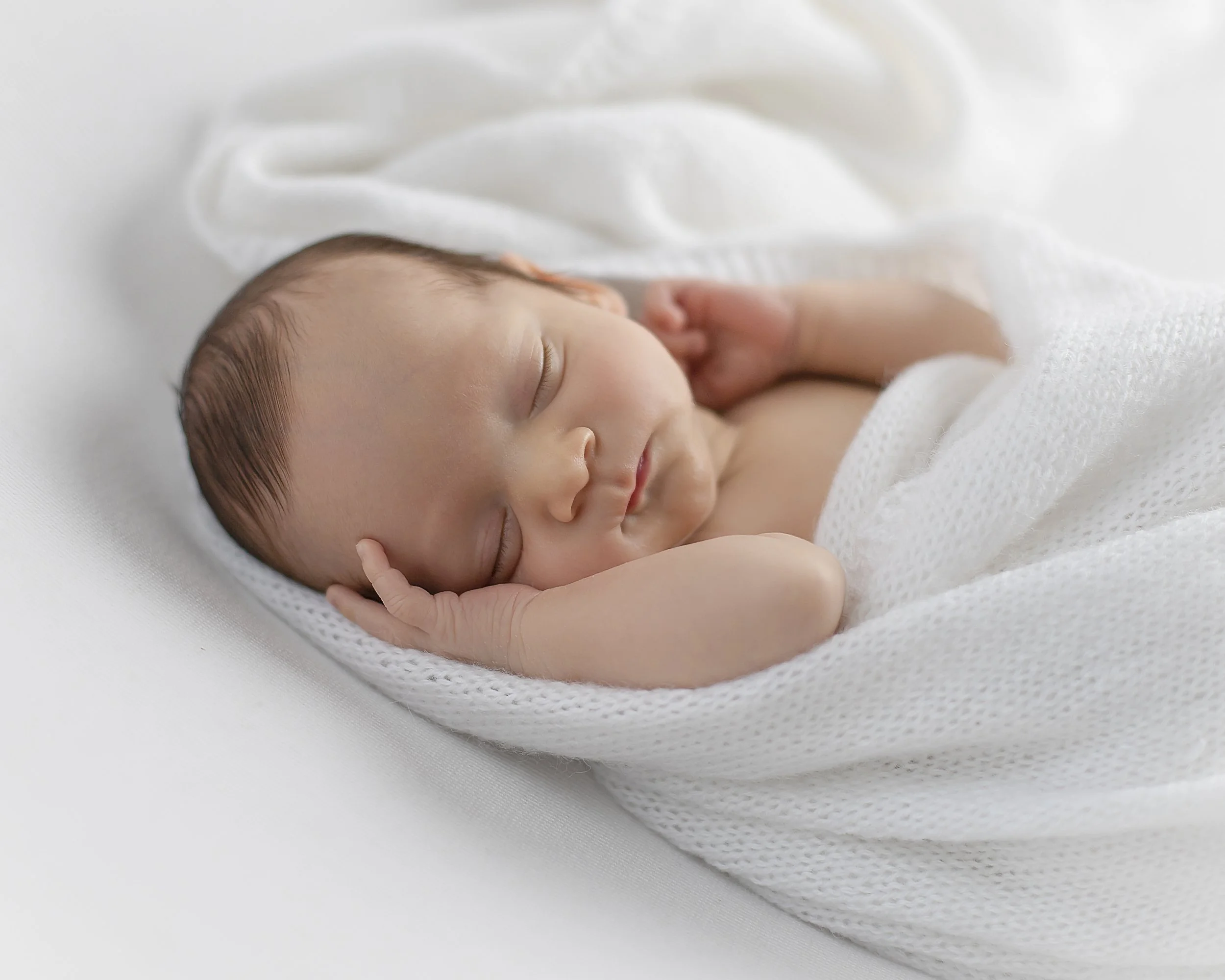 Close-up of a sleeping newborn baby wrapped in a white blanket on a soft white surface.