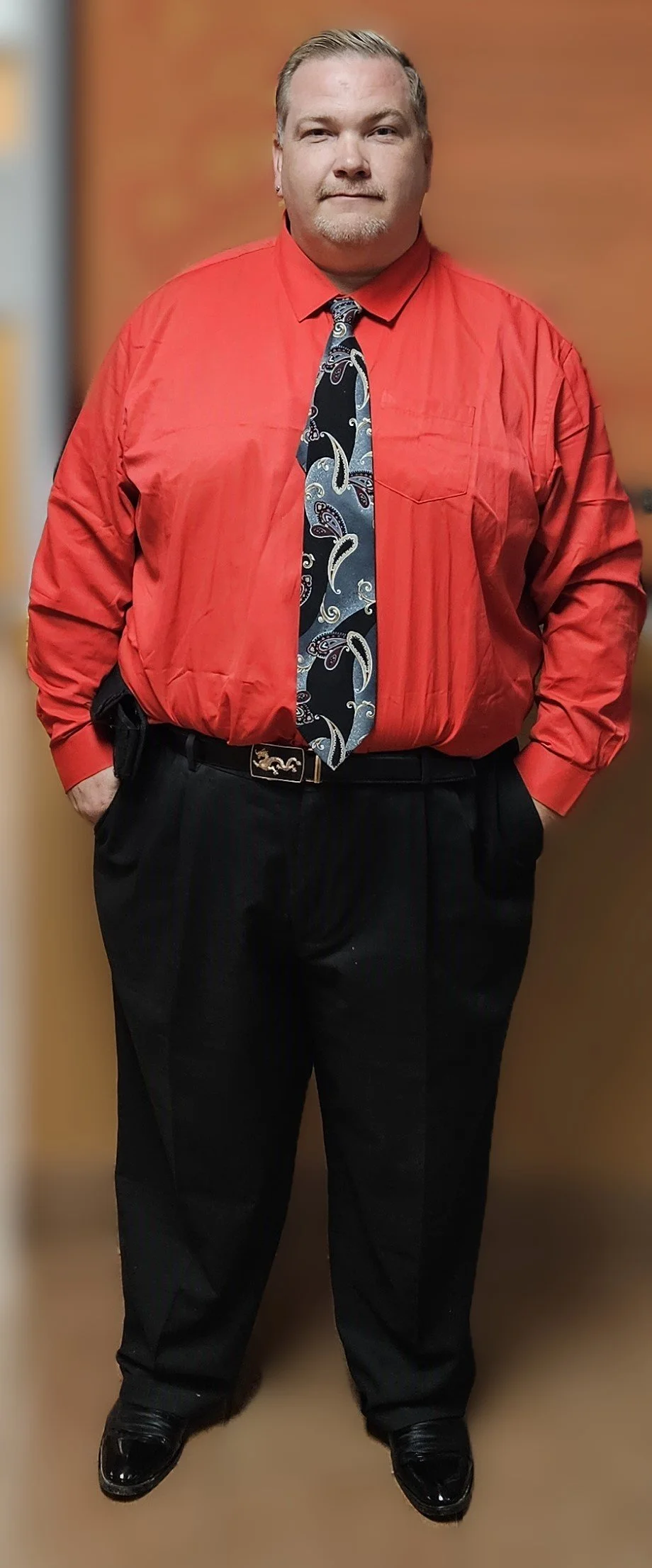 Man wearing a red shirt, paisley tie, and black pants against a neutral background.
