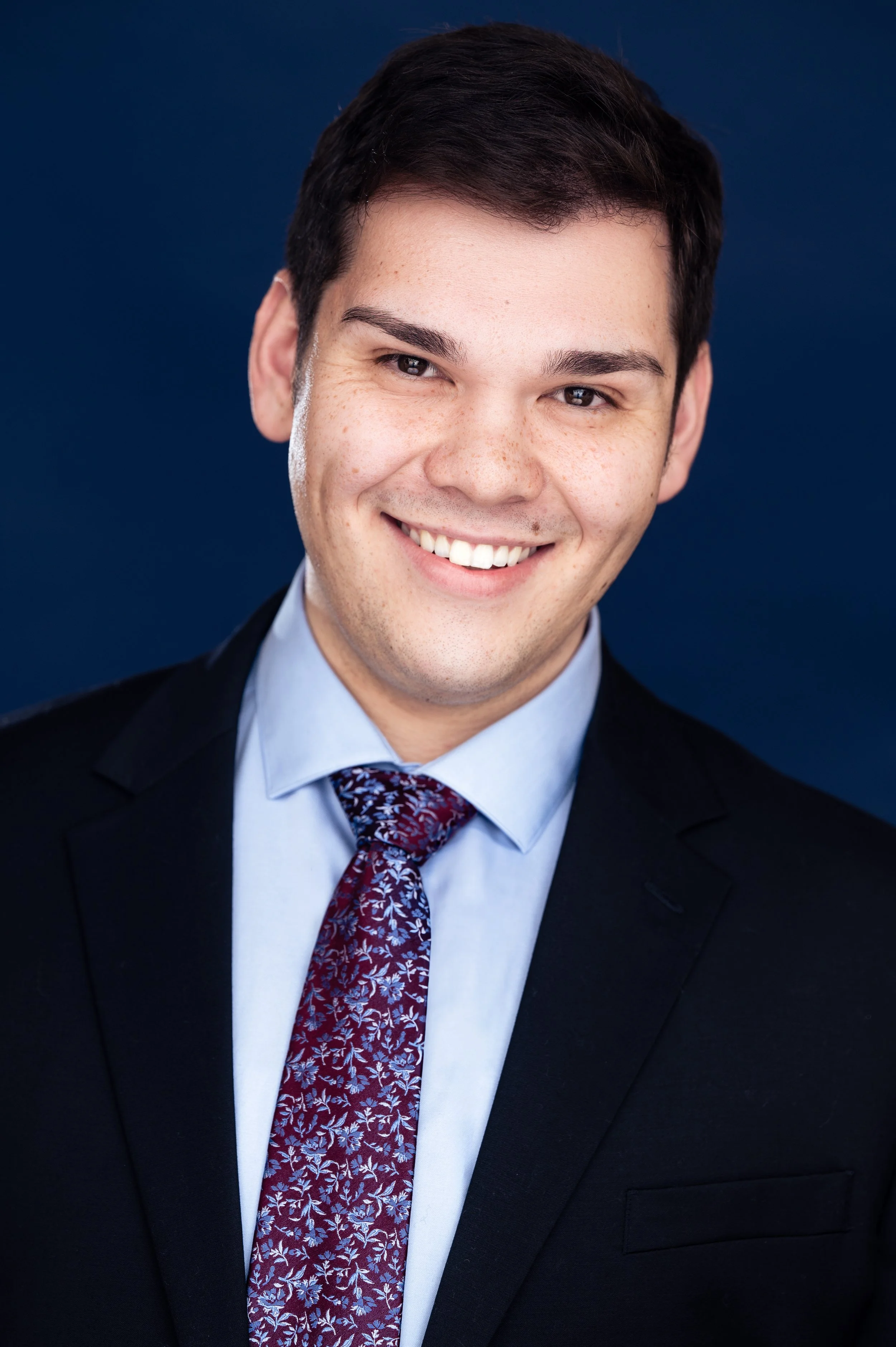 Young man in a suit with a floral tie smiles against a dark blue background.