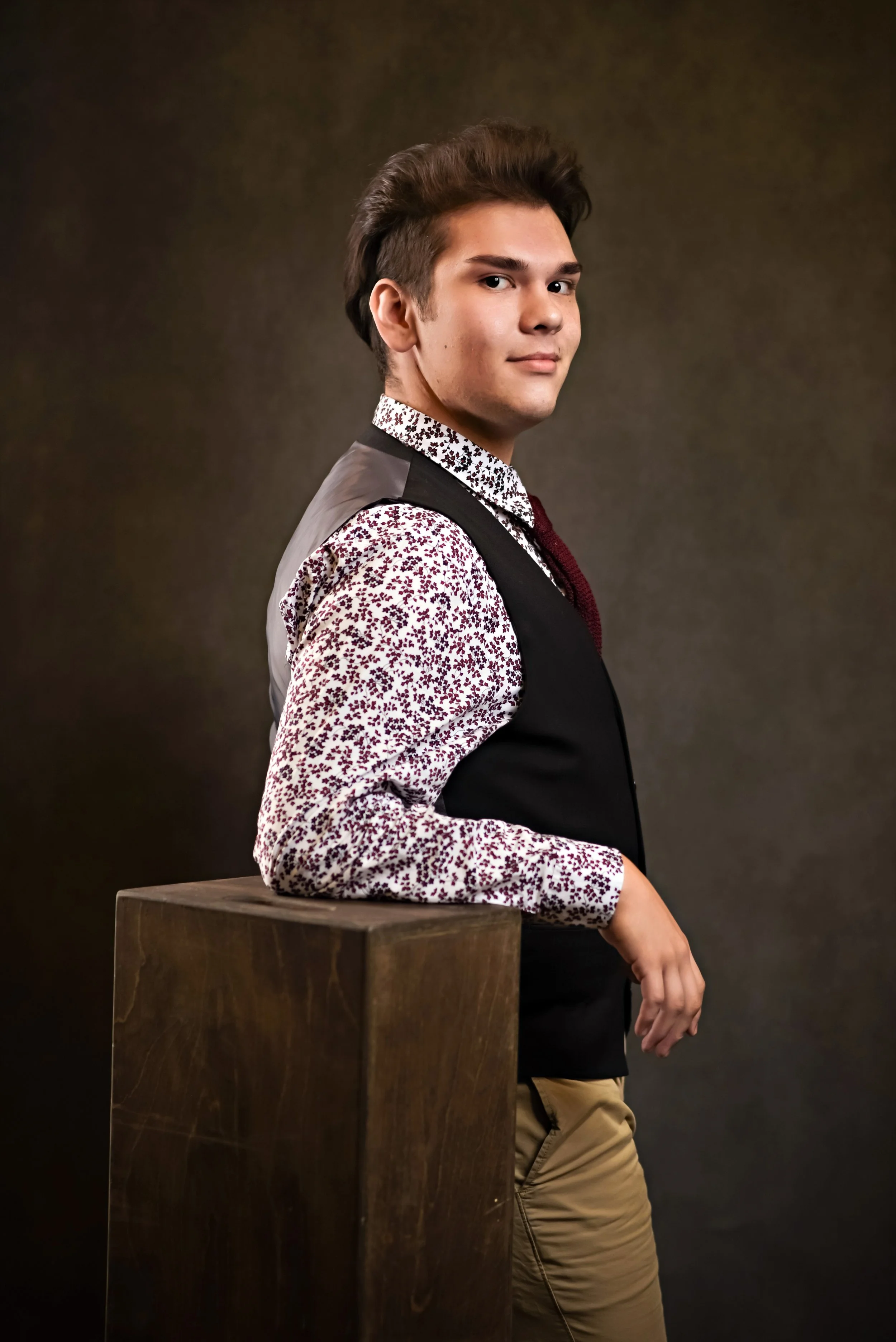 Person wearing a floral shirt, vest, and tie, posing with hand on a wooden block, against a dark background.