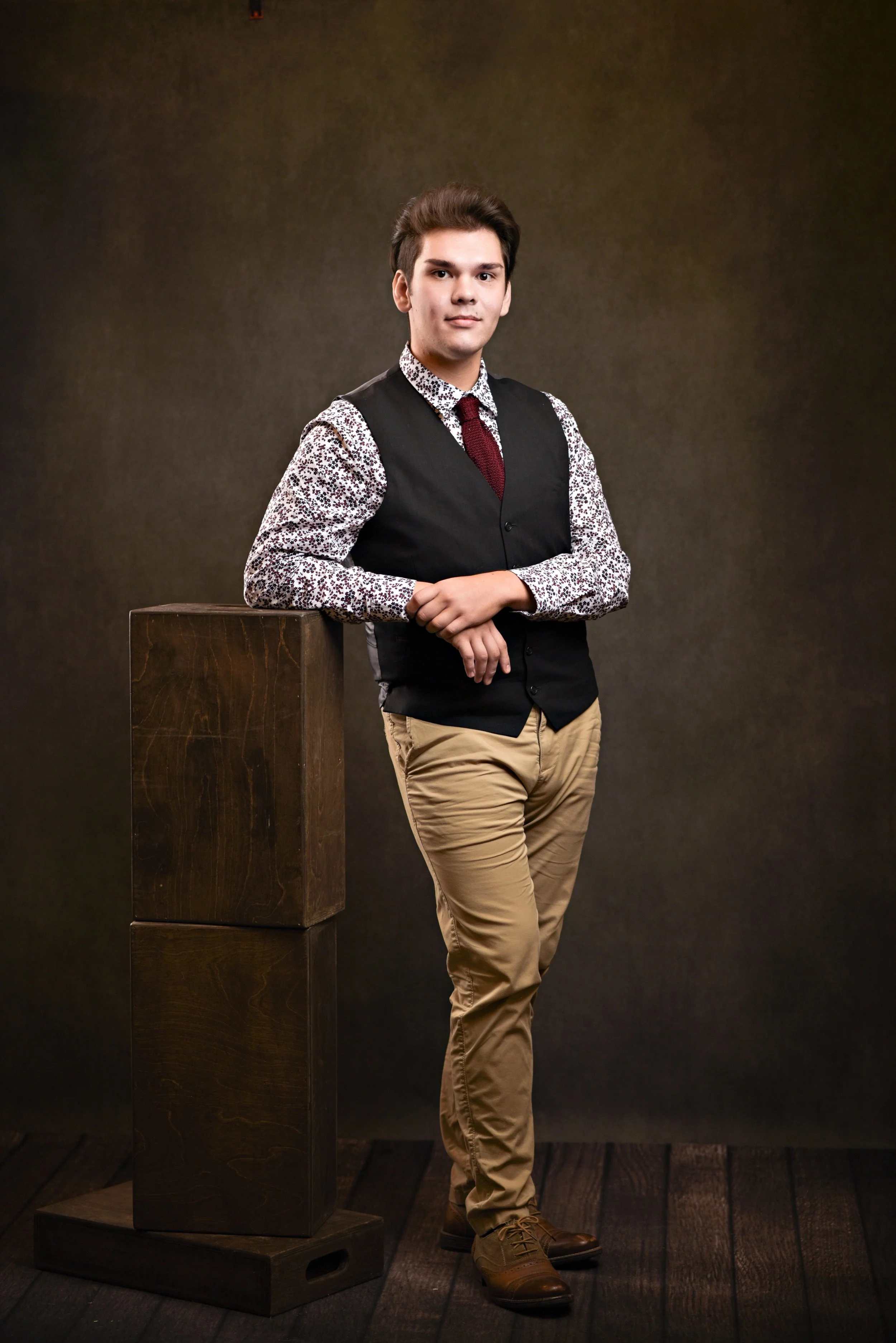 A young man in a floral shirt and black vest stands confidently, leaning on a wooden block, in front of a dark backdrop.