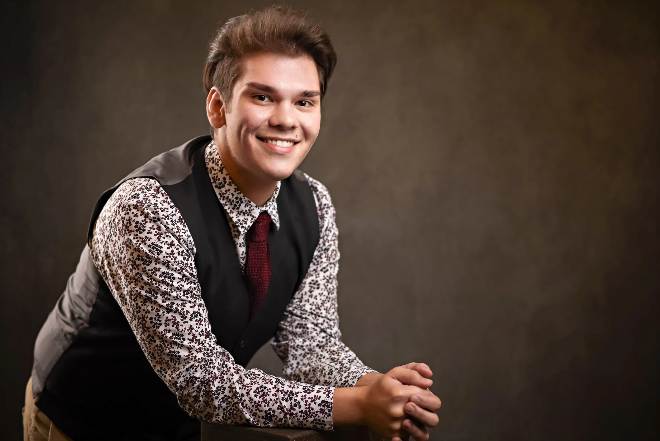 Young man in floral shirt and vest smiling, leaning forward with clasped hands, neutral background.