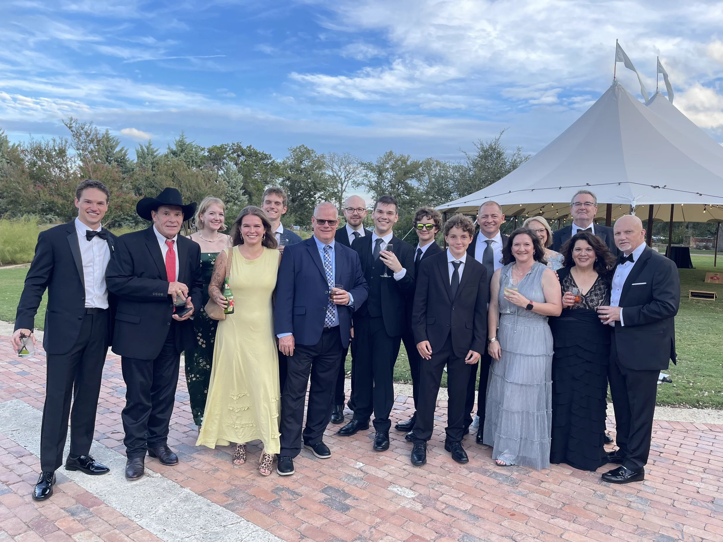 A group of people in formal attire, smiling and posing outdoors in front of a white tent, with a brick pathway and grass. The sky is partly cloudy, and trees are in the background.