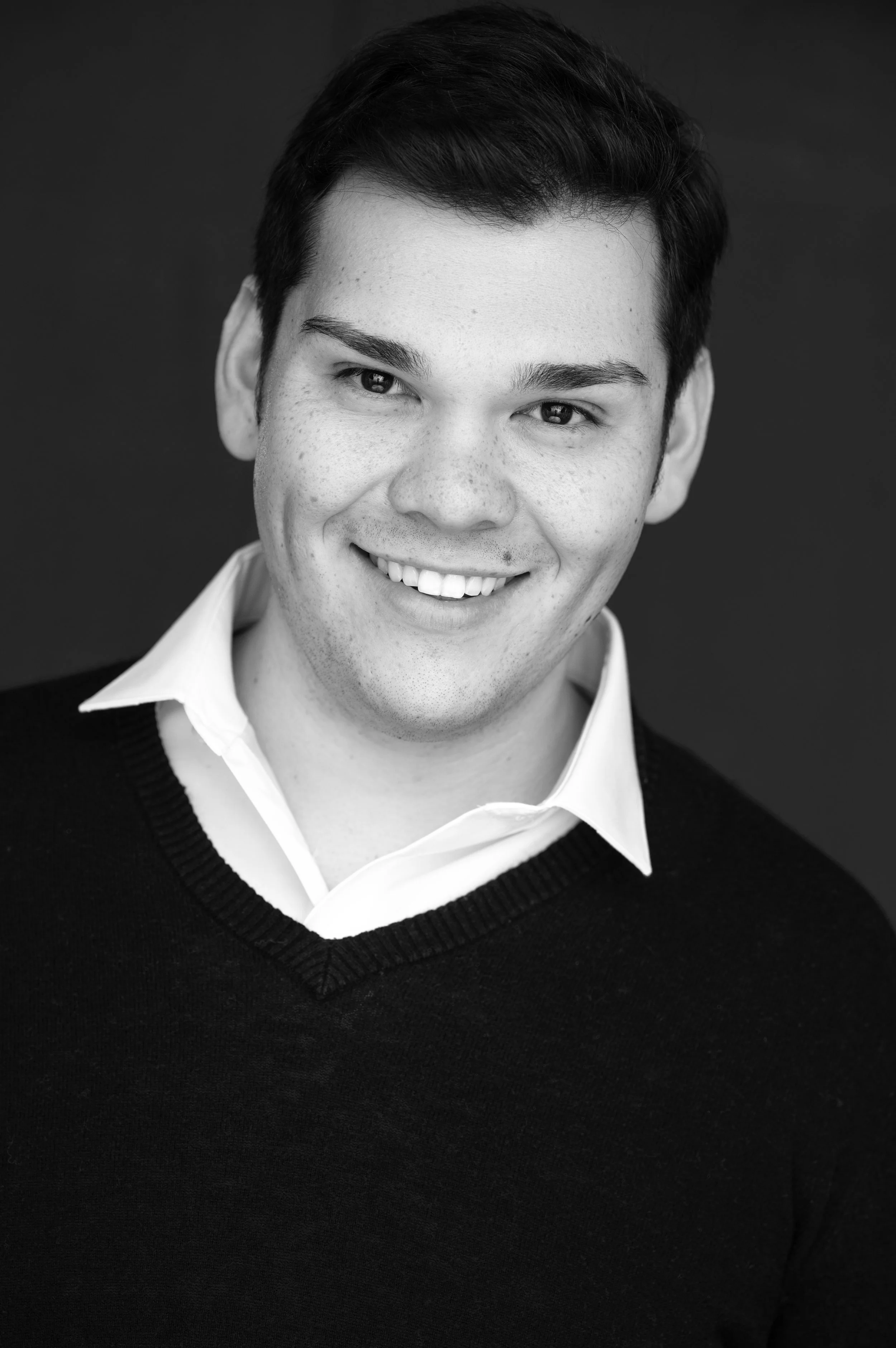 Asa Ambrose, baritone, headshot. Smiling person in a black and white portrait, wearing a white shirt and dark sweater, looking directly at the camera.