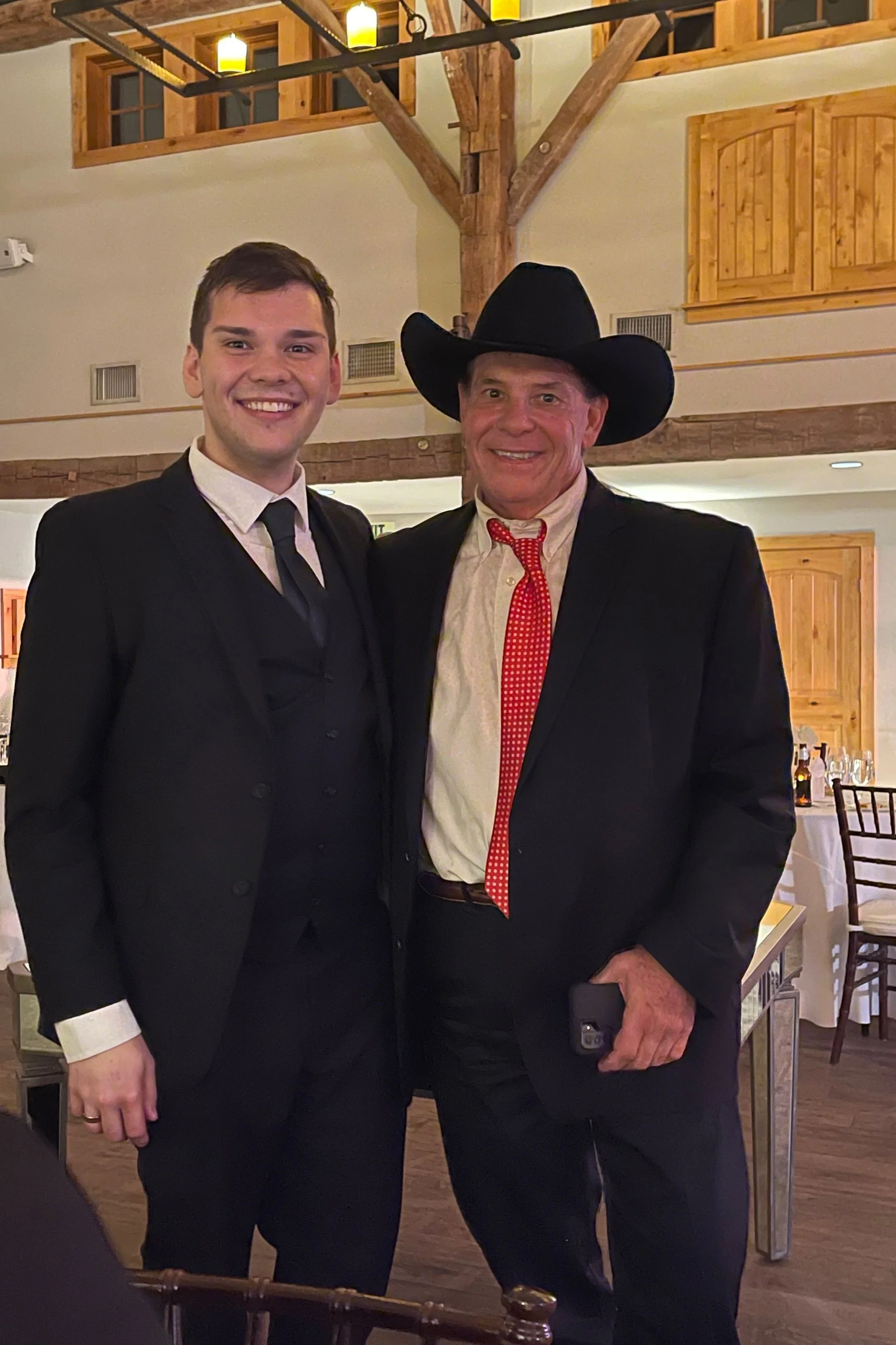 Two men in suits, one wearing a cowboy hat, standing indoors at an event.