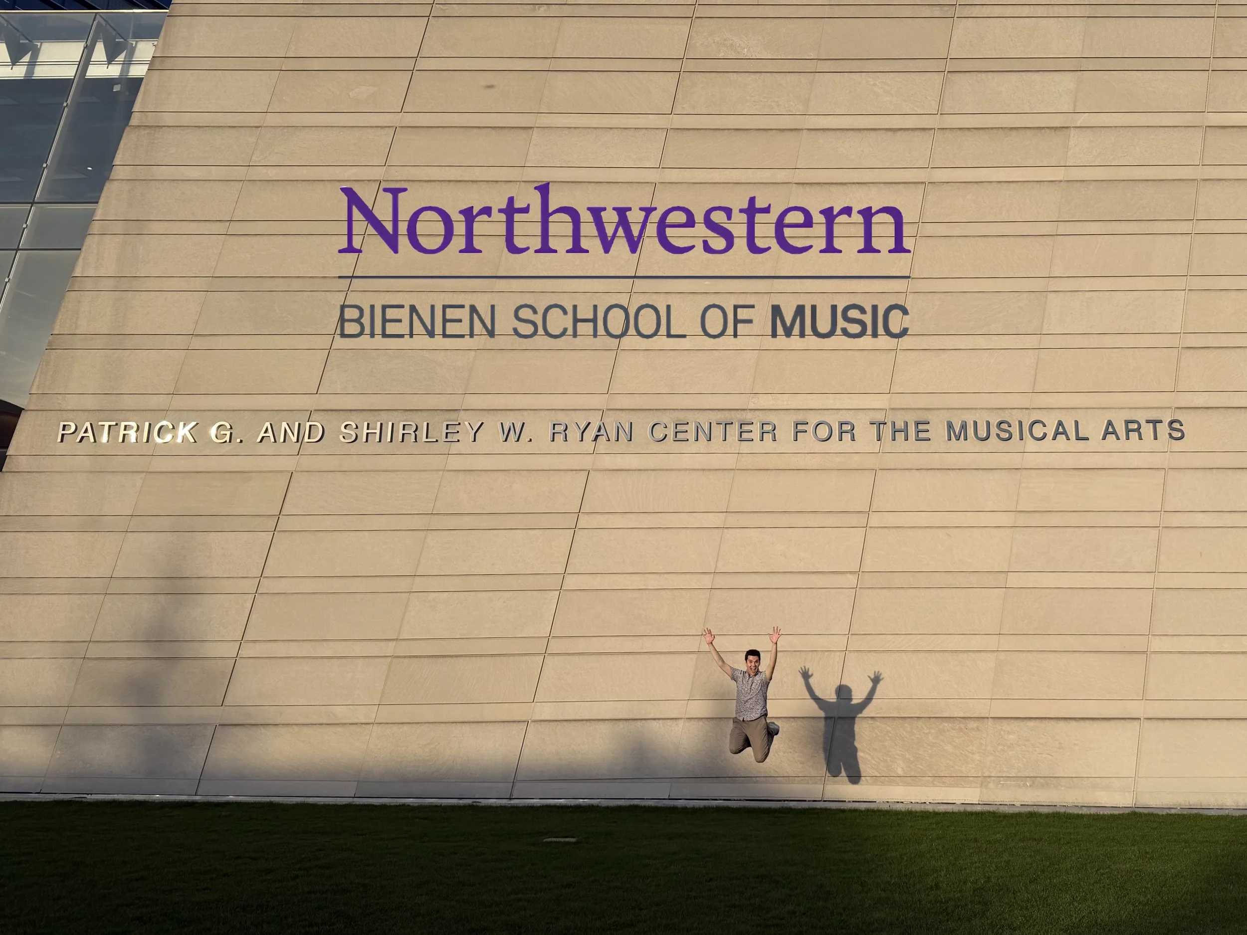 A man is jumping with arms raised in front of Northwestern University's Ryan Center for the Performing Arts with the words 'Northwestern Bienen School of Music' on it.