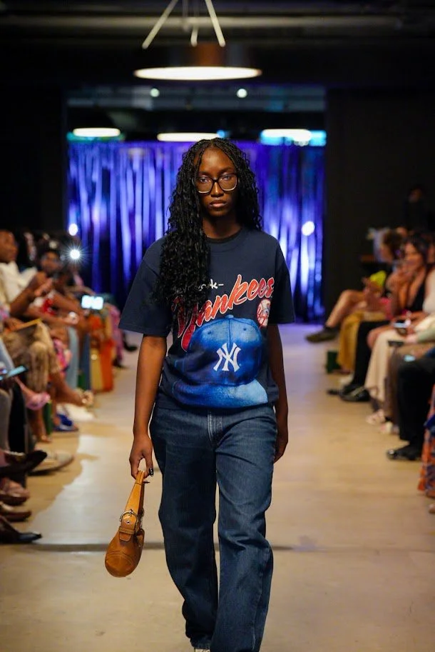 A woman walking down a runway at a fashion show, wearing a New York Yankees T-shirt and blue jeans, holding a small brown handbag, with an audience seated on both sides of the runway and blue stage lighting in the background.