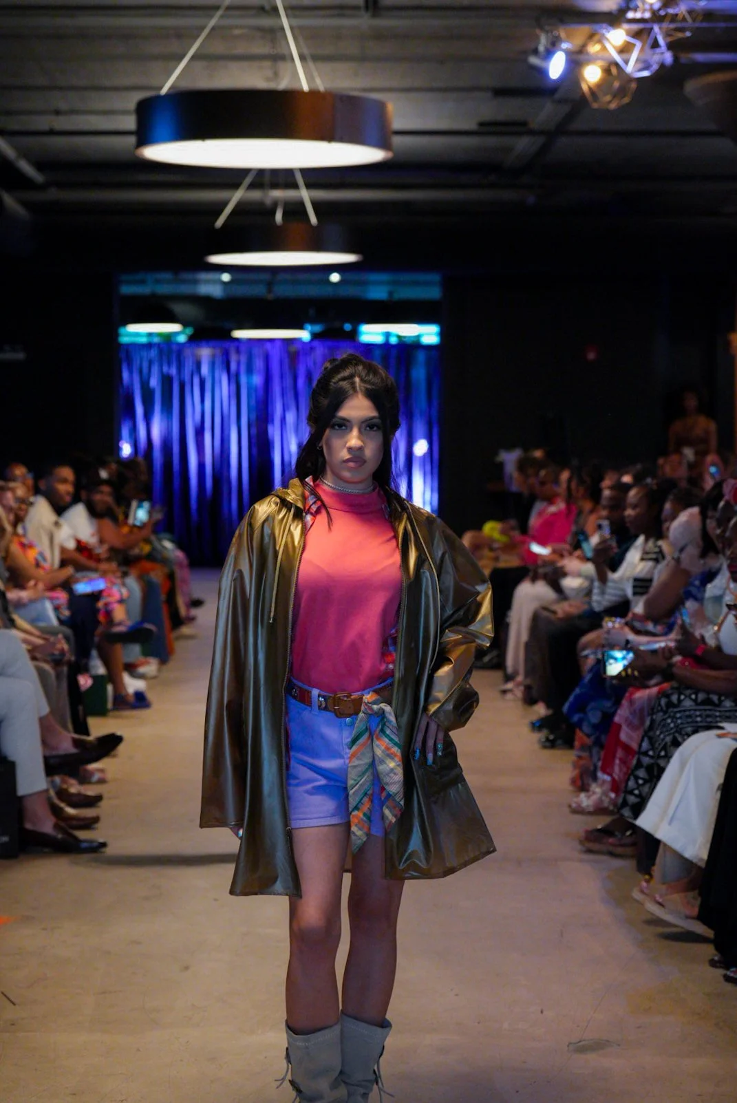 A female runway model walking down the runway during a fashion show, wearing a shiny gold jacket over a pink t-shirt, paired with high-waisted blue shorts and a colorful belt tied in front, in a dimly lit space with an audience on either side.