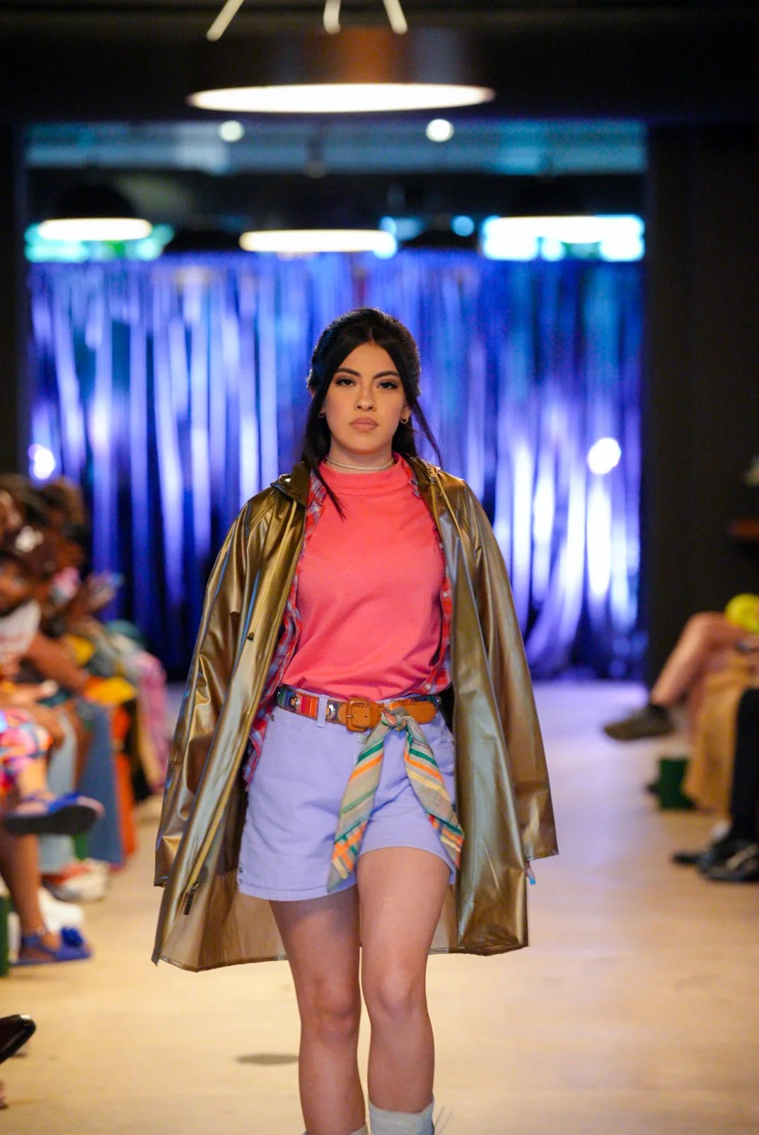 A young woman walks down a runway during a fashion show, wearing a pink t-shirt, lavender shorts with a colorful scarf belt, a brown belt, and a shiny gold jacket. The audience is seated on both sides of the runway, and a blue curtain backdrop is beh