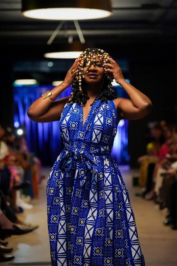 A woman walking on a fashion runway, wearing a sleeveless blue and white patterned dress, with her hands touching her hair styled with beads, and audience members watching in the background.