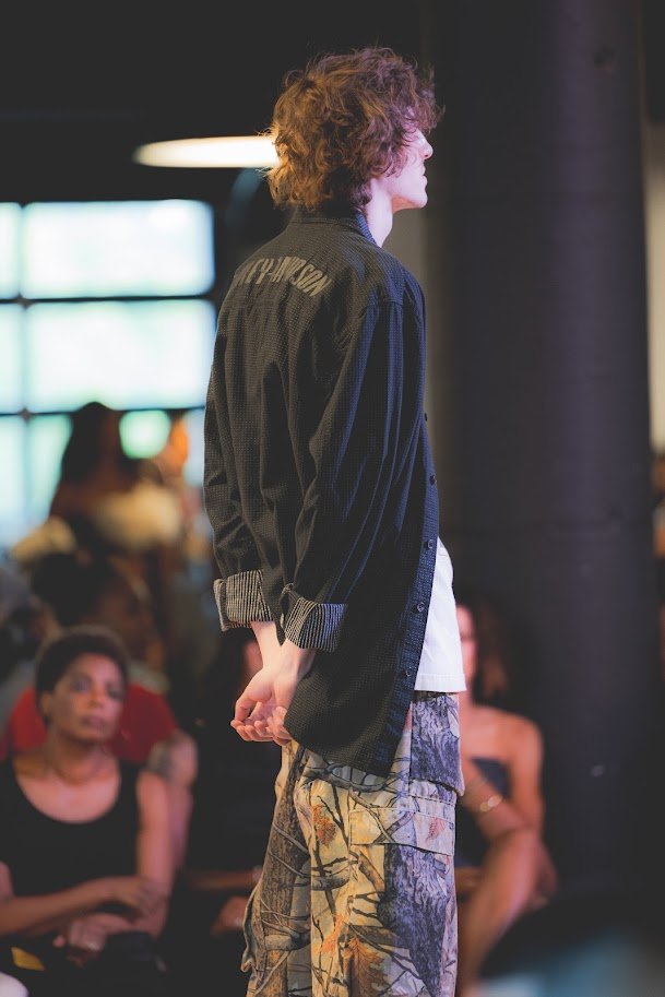 Person with curly hair standing on a runway during a fashion show, wearing camo pants and a dark oversized shirt, with an audience visible in the background.