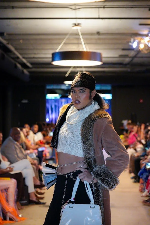 A woman walking on a runway during a fashion show, wearing a leopard print headscarf, a white knit crop top, a faux fur-lined brown coat, black pants with chains, and carrying a white handbag. Audience members are seated on either side of the runway.