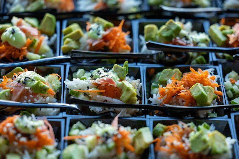 Close-up of individual servings of sushi with rice, sliced avocado, shredded carrots, and black sesame seeds in black plastic containers with black plastic spoons.