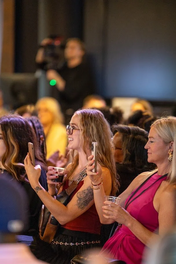 Women in an audience at an event, some taking photos or videos with their phones, smiling and enjoying the moment.