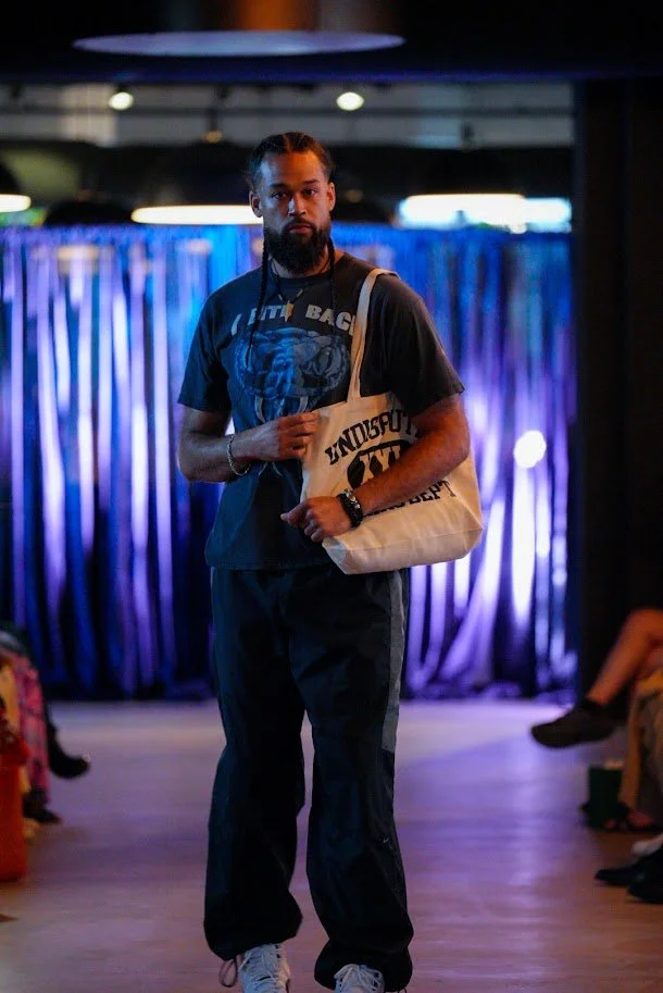 A man with braids and a beard standing on a stage with a blue curtain background, wearing a black T-shirt featuring a graphic, black pants, white shoes, and carrying a beige tote bag with black text.