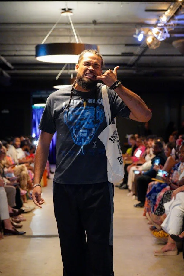 Man smiling and making a gesture with his hand on his face, walking down an aisle at a fashion show with audience seated on both sides.