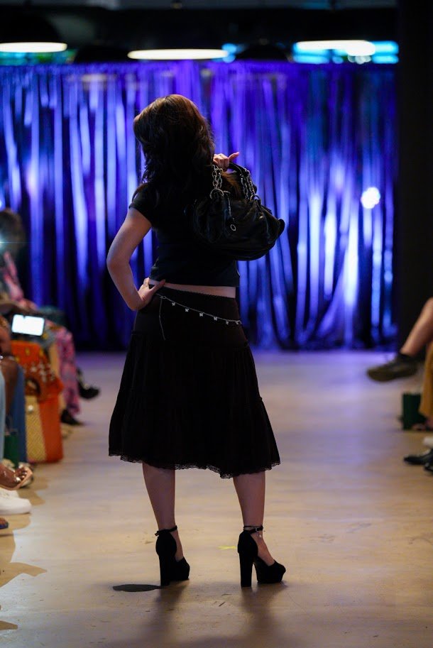 A woman with dark hair modeling on a runway in front of a purple curtain, wearing a black dress and high-heeled shoes, with an audience watching.