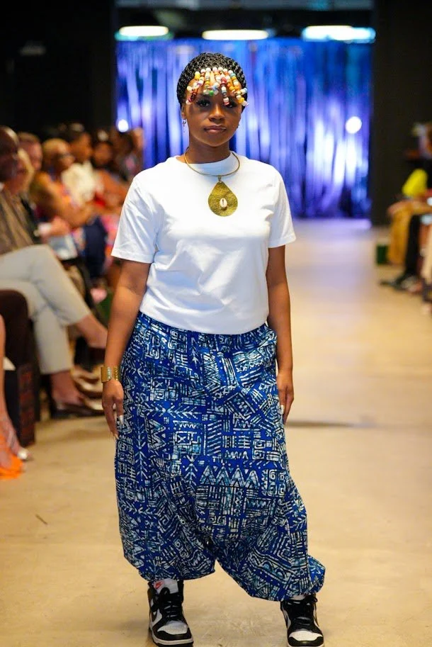 A woman walking on a runway during a fashion show, wearing a white t-shirt, blue patterned pants, and sneakers. She has colorful beaded hair accessories and a large gold pendant necklace.