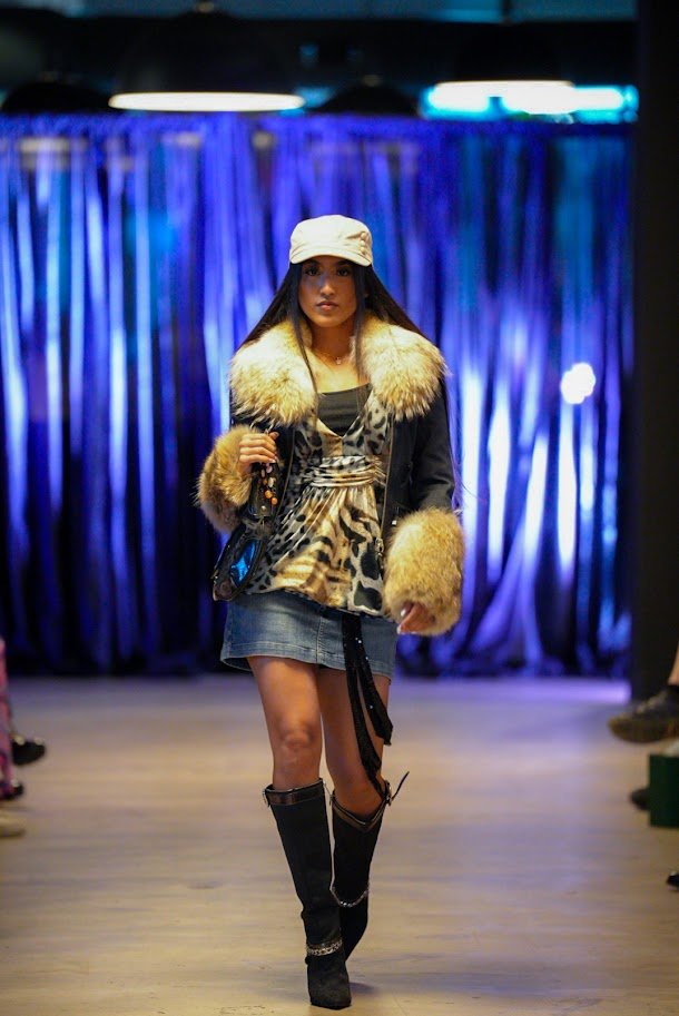 A female model walking on a runway, wearing a beige cap, a fur-lined jacket, a leopard print top, a denim skirt, and black thigh-high boots, with a dark background and blue lighting.