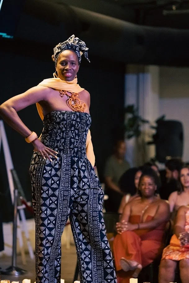 A woman standing confidently on stage, wearing an African-inspired patterned jumpsuit with a yellow scarf draped over her shoulders and a headwrap, with an audience seated and smiling in the background.
