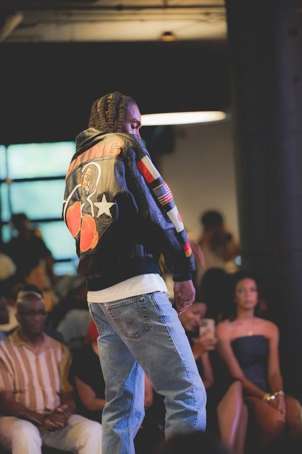 A man with braided hair is walking in front of a seated audience in a dimly lit indoor space. He is wearing a black jacket with colorful patches, including an American flag and a bear graphic, and light blue jeans. The audience members include a woma