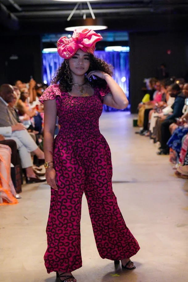 A woman walking on a runway during a fashion show, wearing a pink and black patterned jumpsuit and a large pink headwrap, with fashion audience on either side.
