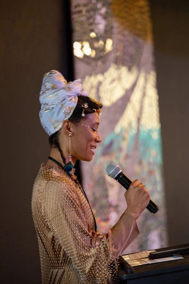 A woman with dark curly hair wrapped in a light-colored head scarf, wearing a beaded top, holding a microphone and standing in front of a colorful projected background.