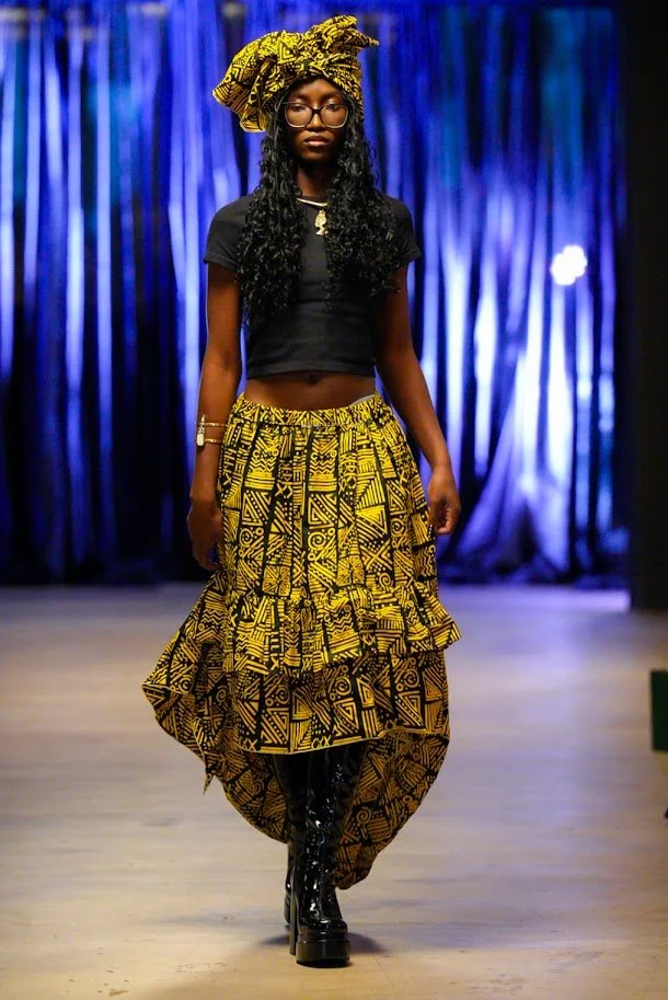 Model walking on runway wearing black crop top and yellow patterned African-inspired skirt and headwrap, with black knee-high boots, against a blue curtain backdrop.