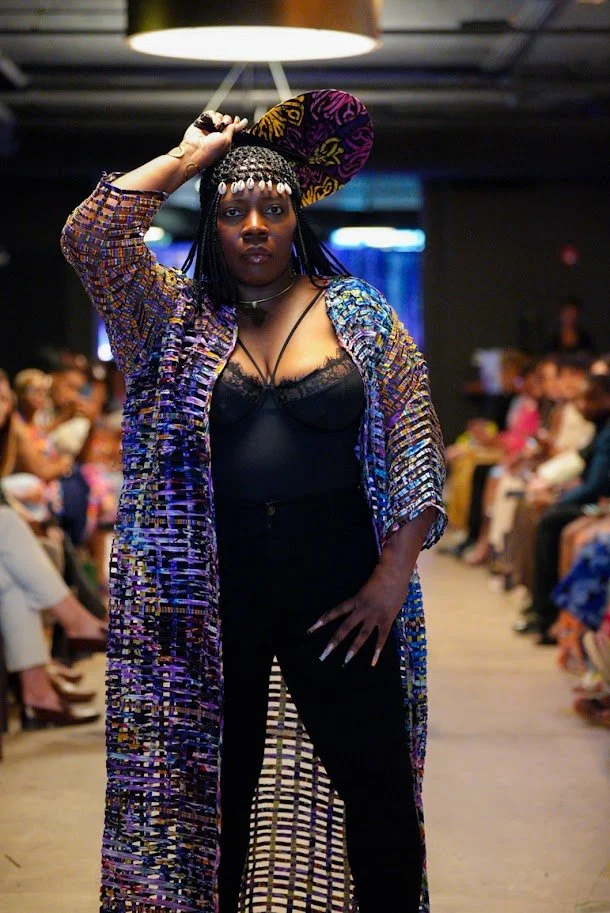 A woman standing confidently on a runway wearing a colorful, patterned long coat, a black lace camisole, black pants, and a decorative hat, with an audience seated on both sides in the background.