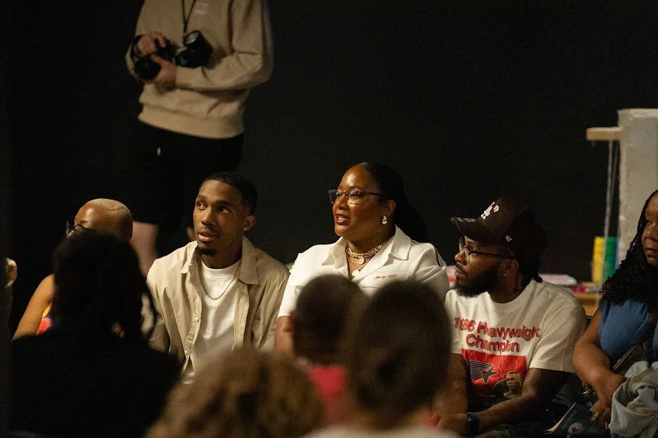 Group of people sitting and listening at an indoor event, some looking towards a speaker or focus, with a person standing in the background holding a camera.