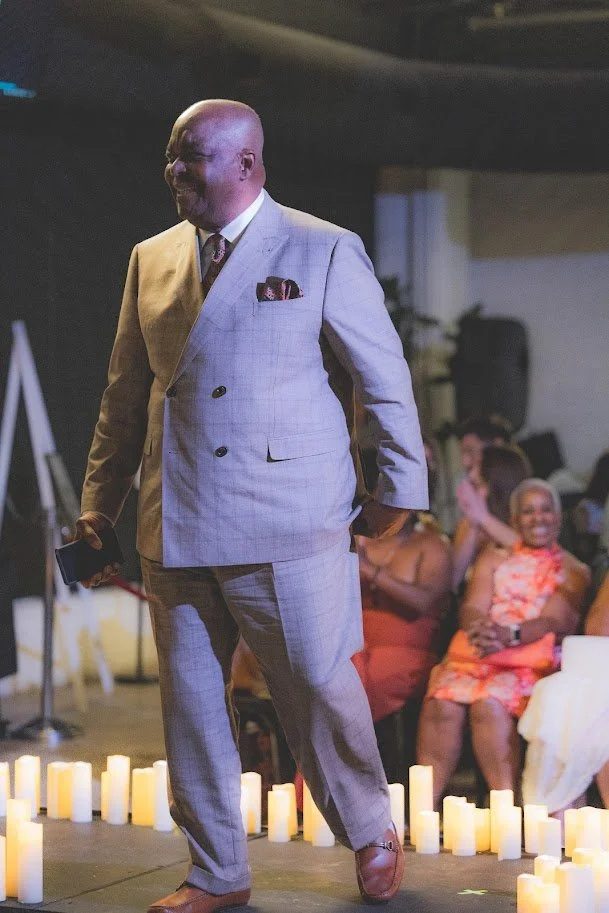 A man in a light gray suit holding a phone standing among battery-operated candles with people sitting and smiling in the background.