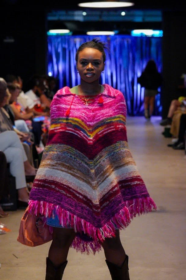 A woman walking on a runway at a fashion show, wearing a colorful, striped poncho with fringe at the bottom. Audience members are seated on either side of the runway, and a dark curtain is in the background.