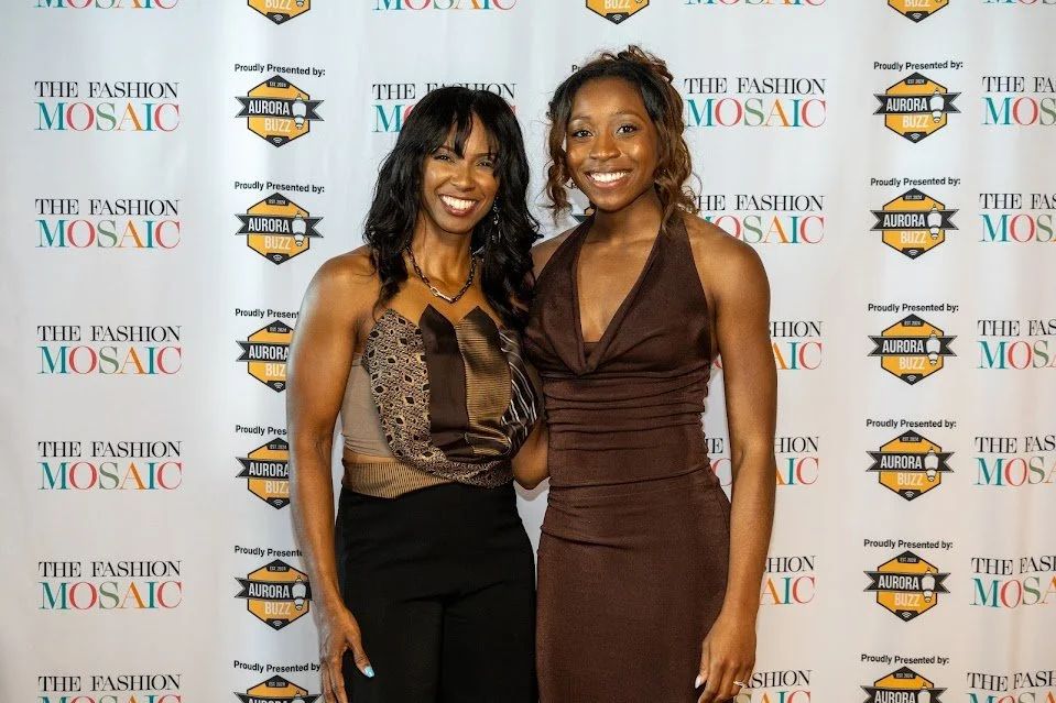 Two women smiling and posing in front of a step and repeat backdrop at a fashion event.