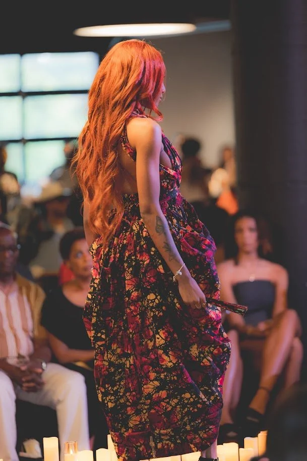 A woman with long, wavy red hair walking on a runway at a fashion show, wearing a colorful, floral dress, holding a phone in her right hand, with an audience watching.