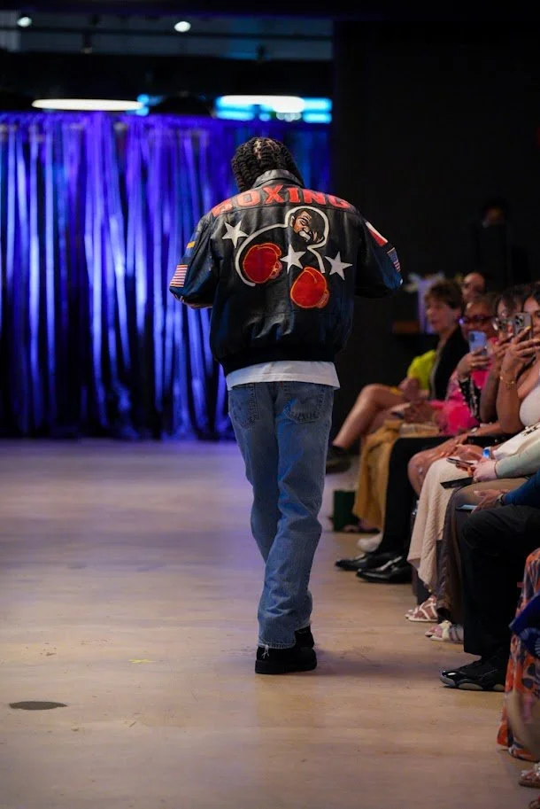 A person walking down a runway at a fashion show wearing a black leather jacket with colorful patches and embroidery, and jeans, with an audience watching on seated on the side.