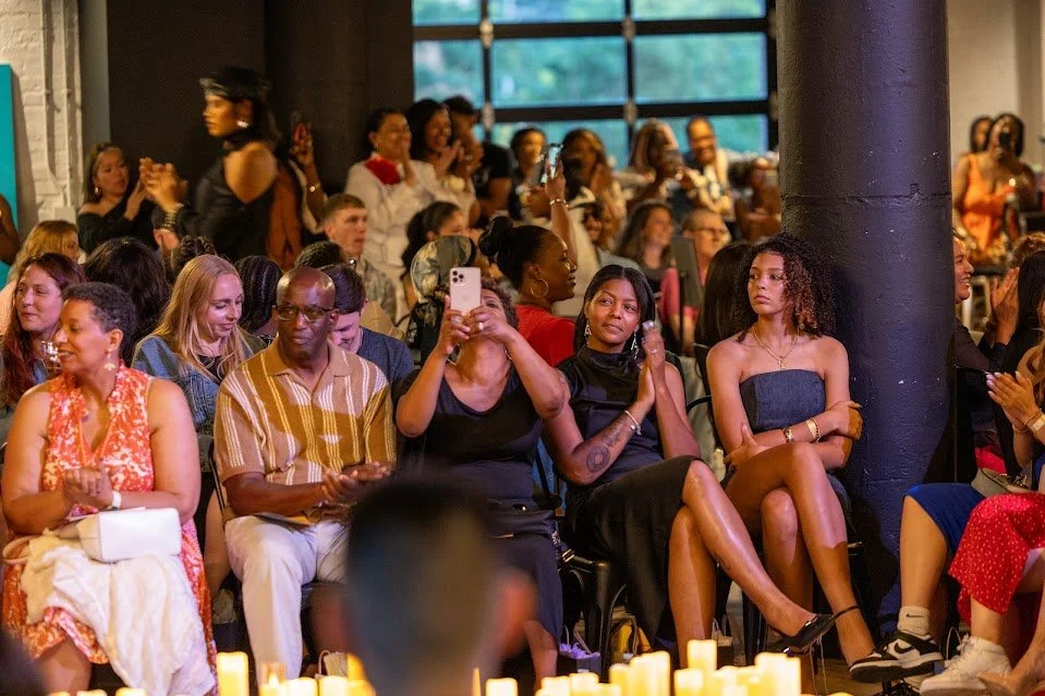 A diverse group of people seated indoors at an event, some clapping, some taking photos, others sitting and watching. There are candles in the foreground and large windows in the background.