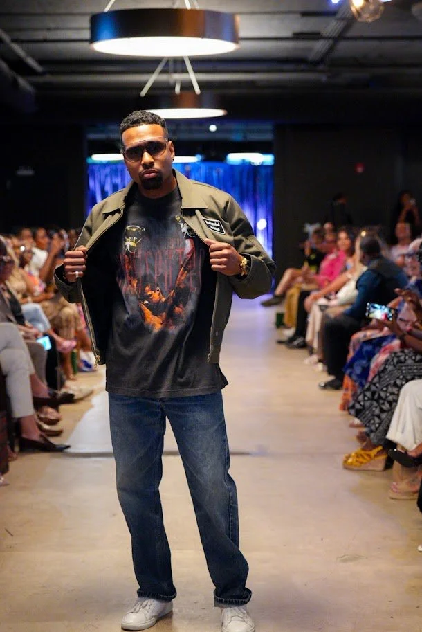 A man walking down a runway during a fashion show, wearing glasses, a green jacket, a graphic t-shirt, jeans, and sneakers, with an audience seated on both sides.