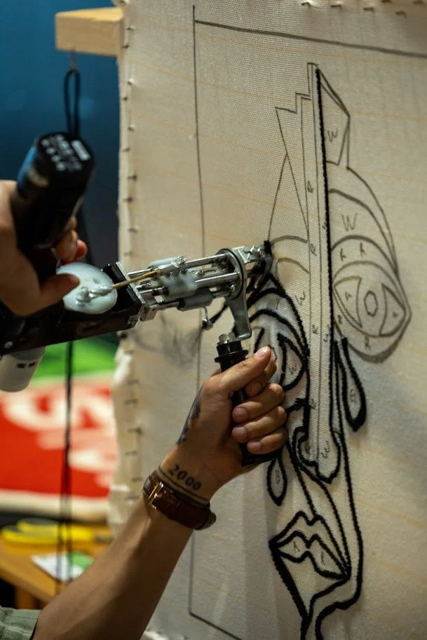 A person is using a tattoo machine to create a large black line drawing of an abstract face on canvas or fabric, with a blue wall and some colorful objects in the background.