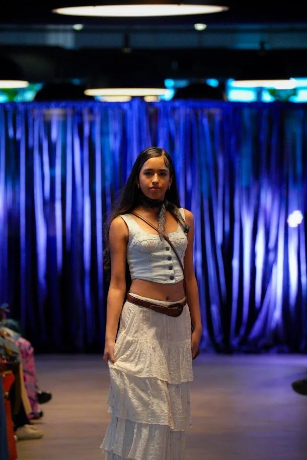 Young woman standing on a stage with a blue curtain backdrop, wearing a light blue sleeveless crop top, white layered skirt, and a dark belt, in a setting with audience members visible on the sides.