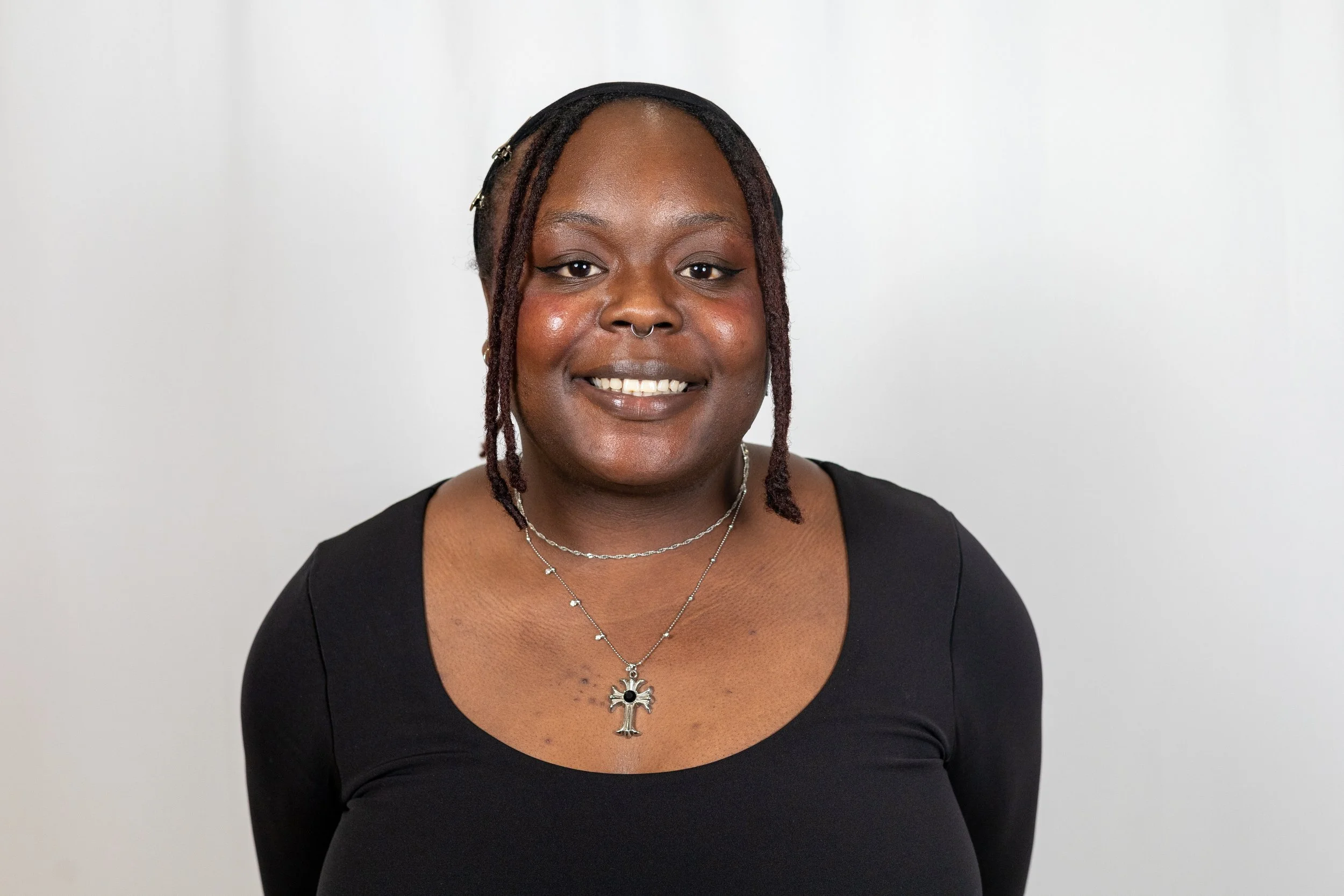 A woman with dark skin and dreadlocks wearing a black top, silver jewelry, a septum piercing, and a big smile against a plain white background.