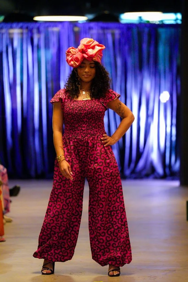 Woman on a runway wearing a pink animal print jumpsuit and a large pink headwrap, with a blue curtain backdrop.