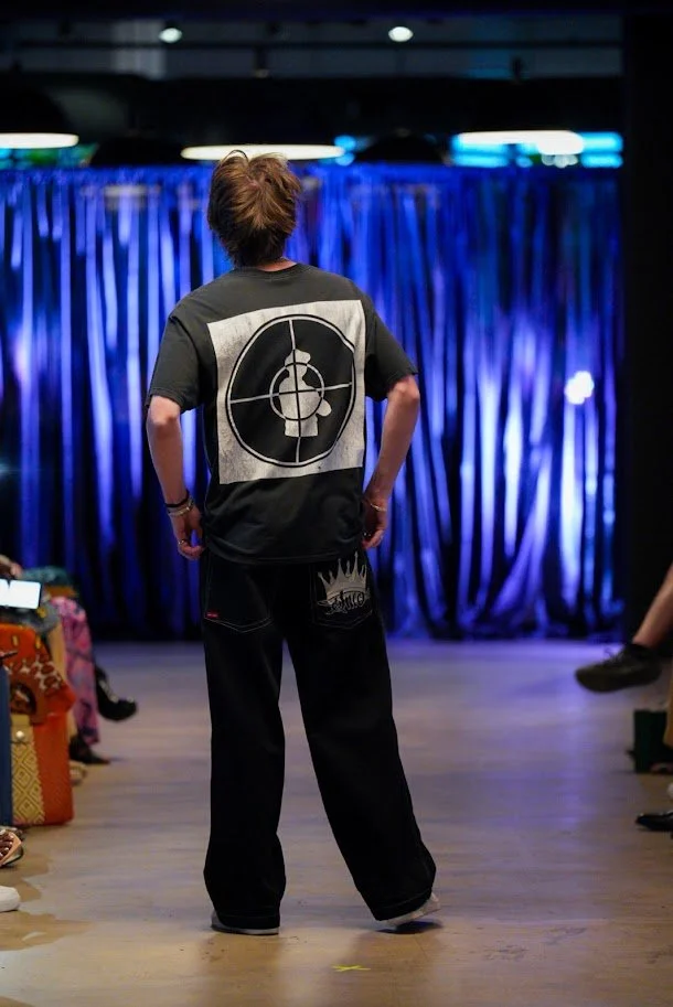 Person with short hair wearing a black t-shirt with a beheaded skull target design on the back, and black pants with a graphic near the bottom, standing on a stage with a blue curtain backdrop.