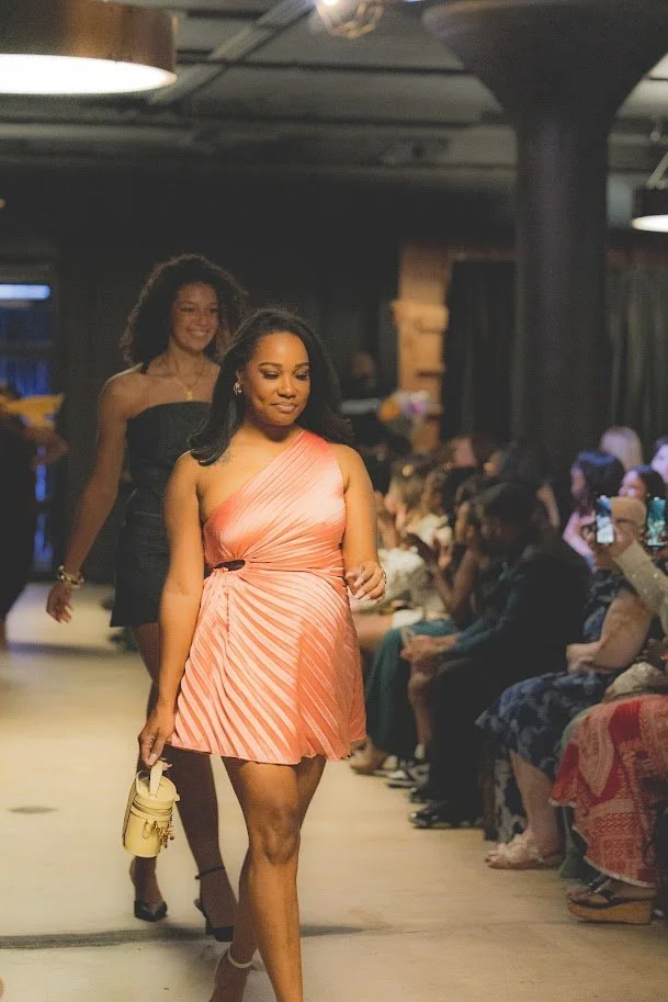 A woman in a peach-colored, striped dress walks on a runway during a fashion show, with an audience seated on either side watching and some taking photos.