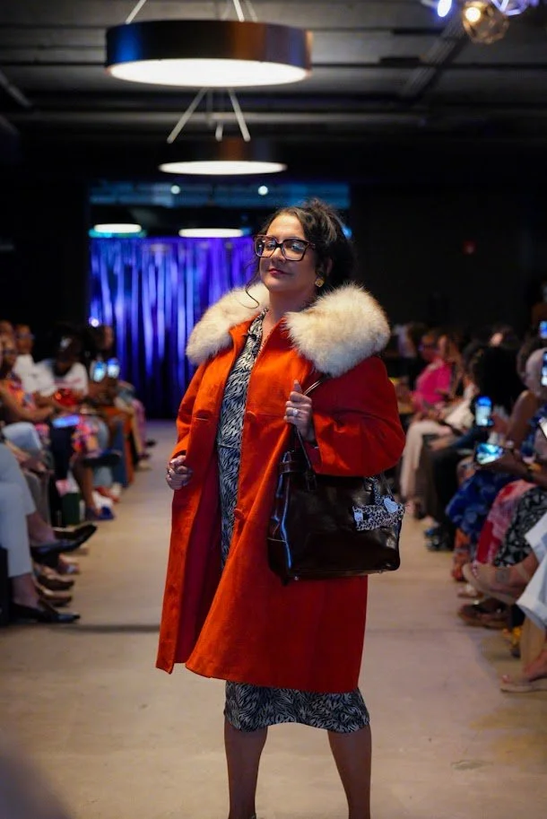 Woman modeling fashion on runway, wearing an orange coat with fur collar, patterned dress, glasses, and carrying a purse, with seated audience along the sides.